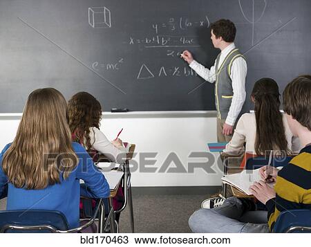 Stock Image - Teacher writing on chalkboard in classroom. Fotosearch