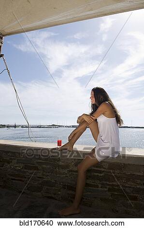 Stock Image - Woman sitting on wall overlooking ocean. Fotosearch