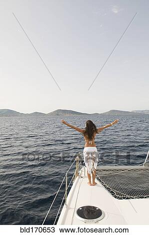 Stock Image - Woman standing with arms outstretched on sailboat deck. Fotosearch