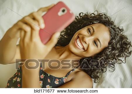 Stock Photo - Woman taking selfie with cell phone on bed. Fotosearch