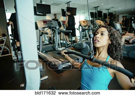 Stock Image - Woman using exercise machine in gymnasium. Fotosearch