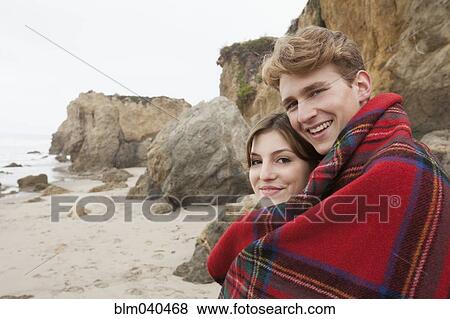 Stock Photo - Caucasian couple wrapped in blanket on beach. Fotosearch