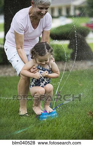Stock Photo - Caucasian grandmother and granddaughter playing in sprinkler. Fotosearch