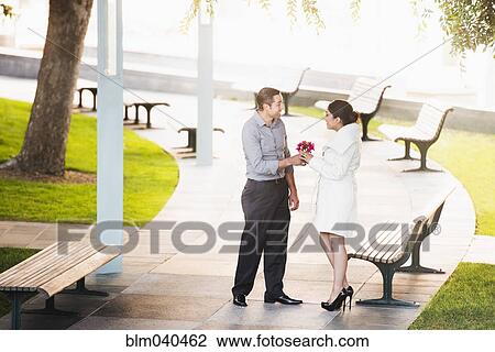Stock Image - Hispanic man giving girlfriend flowers in park. Fotosearch