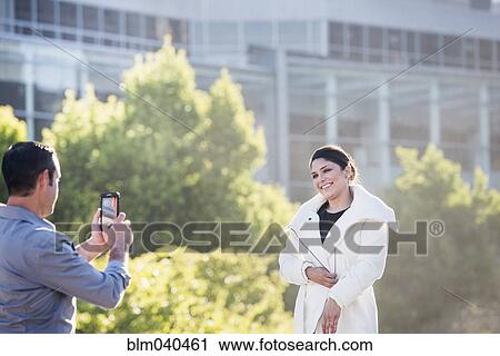 Stock Image - Hispanic man taking cell phone picture of girlfriend in city. Fotosearch