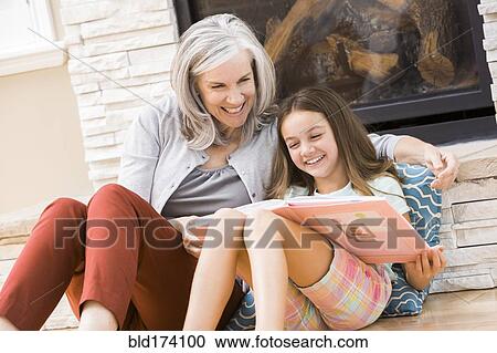 Stock Image - Caucasian grandmother and granddaughter reading book by fireplace. Fotosearch