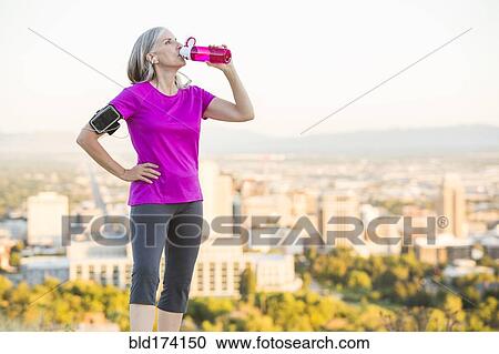 Stock Image - Caucasian woman drinking water bottle on urban hilltop. Fotosearch