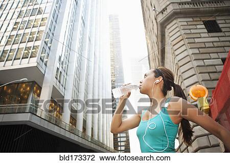 Stock Image - Hispanic woman drinking water bottle under highrise buildings. Fotosearch