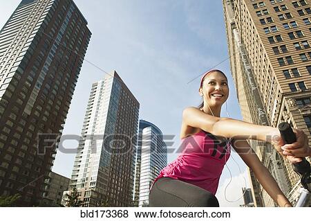 Stock Photo - Hispanic woman riding bicycle under highrise buildings. Fotosearch