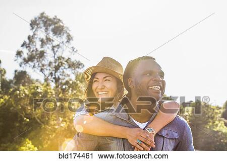 Stock Image - Man carrying girlfriend piggyback outdoors. Fotosearch