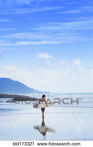 Stock Image - Man carrying surfboard on beach. Fotosearch