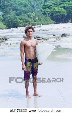 Stock Image - Man carrying surfboard on beach. Fotosearch
