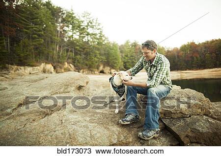 Stock Image - Man petting dog on rocks near lake. Fotosearch