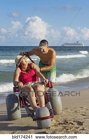 Stock Image - Man pushing paraplegic girlfriend in wheelchair on beach. Fotosearch