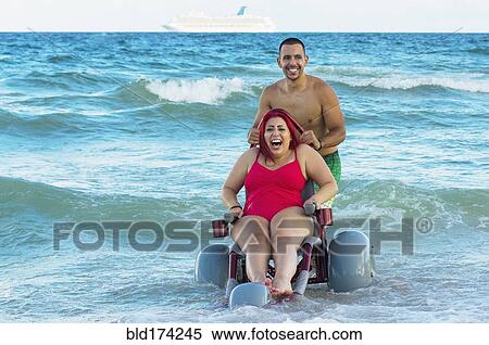 Stock Photography - Man pushing paraplegic girlfriend in wheelchair on beach. Fotosearch