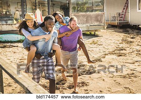 Stock Photo - Men carrying girlfriends piggyback in sand. Fotosearch