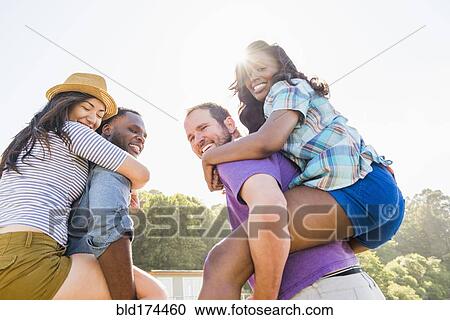 Stock Image - Men carrying girlfriends piggyback outdoors. Fotosearch