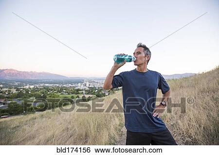 Stock Photograph - Mixed race man drinking water bottle on hilltop. Fotosearch