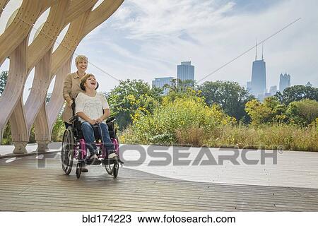 Stock Image - Mother pushing paraplegic daughter in wheelchair. Fotosearch