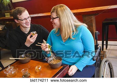 Stock Image - Paraplegic woman and boyfriend eating noodles in tea shop. Fotosearch