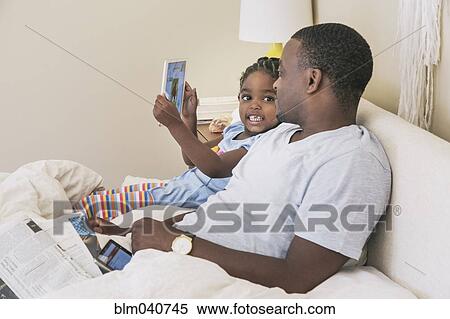Stock Photography - African American father and daughter using digital tablet on bed. Fotosearch