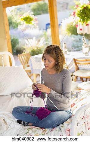 Stock Image - Mixed race woman knitting on bed. Fotosearch