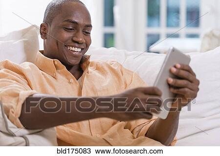 Stock Image - Black man using digital tablet on sofa. Fotosearch