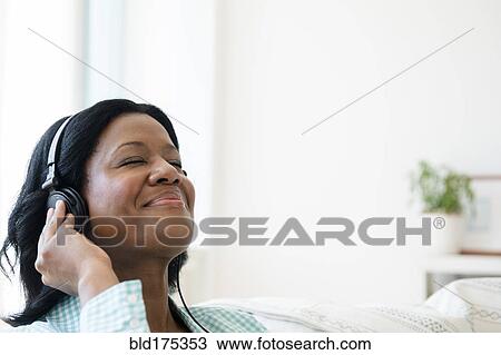 Stock Image - Black woman listening to headphones on sofa. Fotosearch