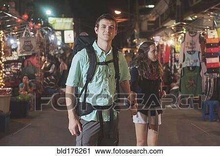 Stock Image - Caucasian tourists holding hands in market at night. Fotosearch