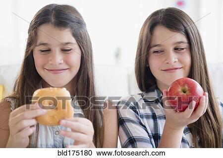 Stock Image - Caucasian twin sisters eating healthy and unhealthy snacks. Fotosearch