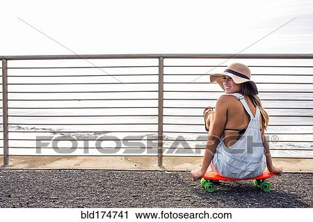Stock Image - Caucasian woman sitting on skateboard at beach. Fotosearch
