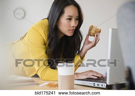 Stock Image - Chinese businesswoman using laptop and eating. Fotosearch