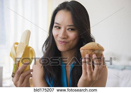 Stock Photo - Chinese woman choosing between banana and muffin. Fotosearch