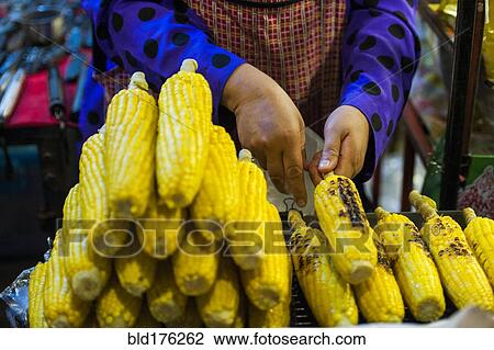 Stock Image - Close up of seller grilling corn at market. Fotosearch