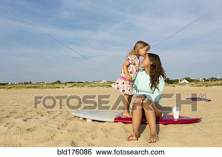 Stock Photograph - Girl kissing forehead of mother on surfboard on beach. Fotosearch