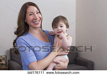 Stock Image - Mixed race mother holding baby daughter in living room. Fotosearch