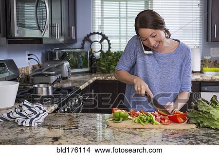 Picture - Mixed race woman talking on phone and chopping vegetables. Fotosearch