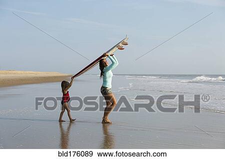 Stock Photo - Mother and daughter carrying surfboard on beach. Fotosearch