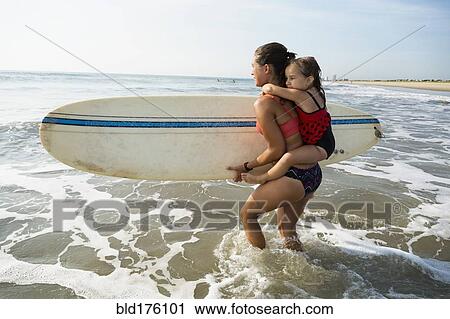 Stock Image - Mother carrying daughter and surfboard on beach. Fotosearch