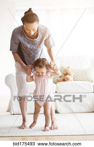 Stock Image - Mother helping baby daughter walk in living room. Fotosearch