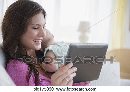 Stock Image - Mother using digital tablet and holding baby daughter. Fotosearch