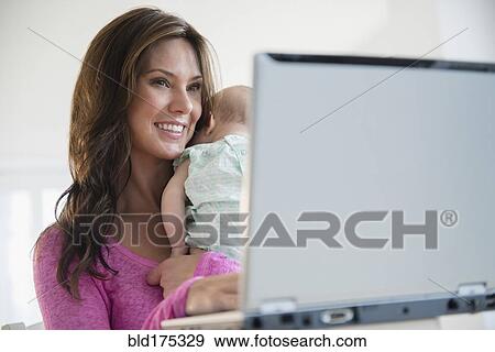 Stock Photo - Mother using laptop and holding baby daughter. Fotosearch