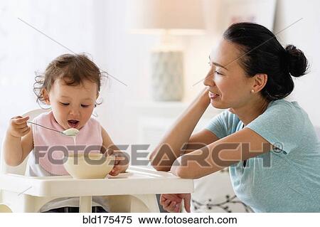 Stock Photography - Mother watching baby daughter eat in high chair. Fotosearch