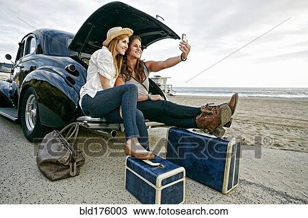 Stock Image - Women with luggage and vintage car on beach. Fotosearch