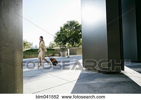 Stock Image - Asian businesswoman rolling luggage outdoors. Fotosearch