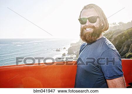 Stock Photo - Caucasian man holding surfboard on coastal cliff. Fotosearch