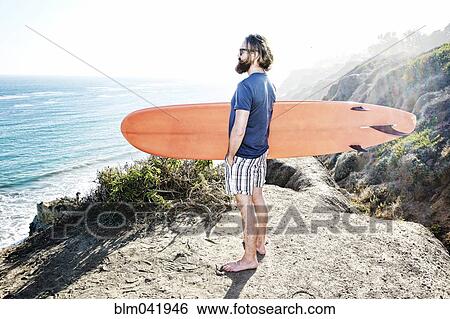 Stock Photograph - Caucasian man holding surfboard on coastal hilltop. Fotosearch