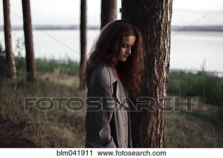 Stock Image - Caucasian woman standing behind tree near rural river. Fotosearch