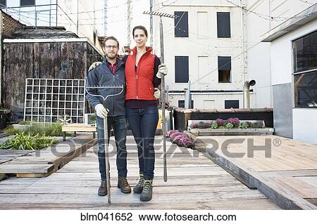 Stock Image - Couple holding rakes in urban rooftop garden. Fotosearch