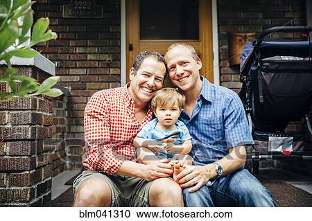 Stock Image - Gay fathers holding baby son on front stoop. Fotosearch